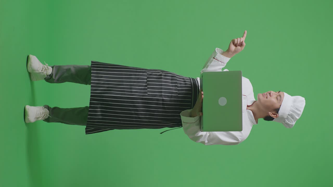 Full Body Of Asian Man Chef Using A Laptop And Pointing To Side While Standing In The Green Screen Background Studio