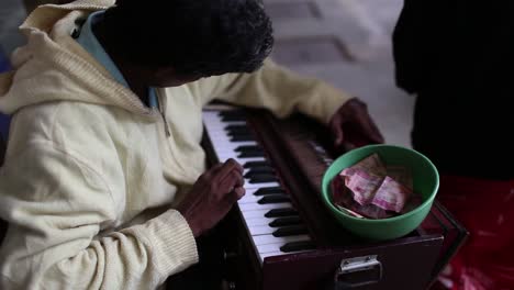 blind indian street artist plays traditional piano on the sidewalk, collecting rupees in the pot