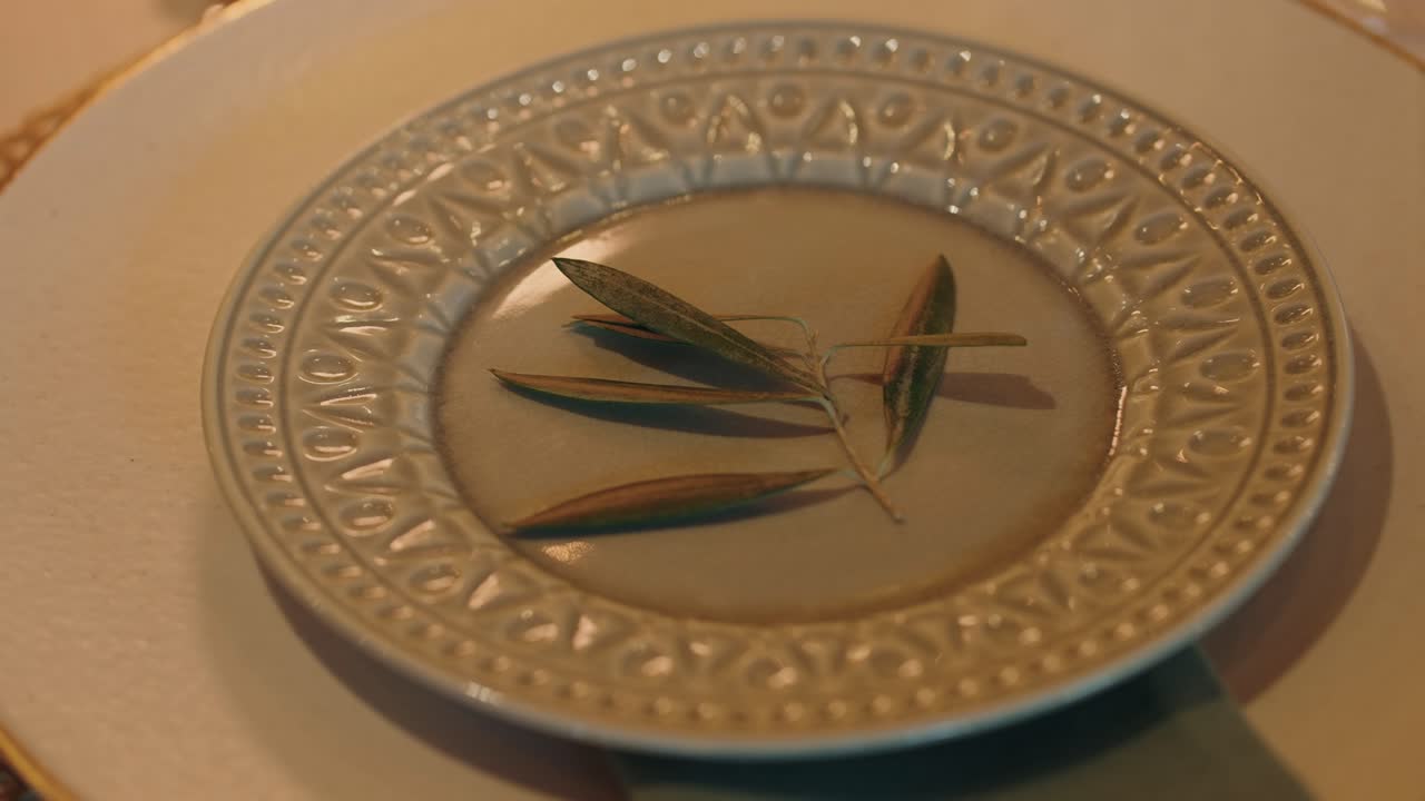 Dettaglio raffinato di un tavolo nuziale con piatto decorato e centrotavola con rametto d'ulivo.