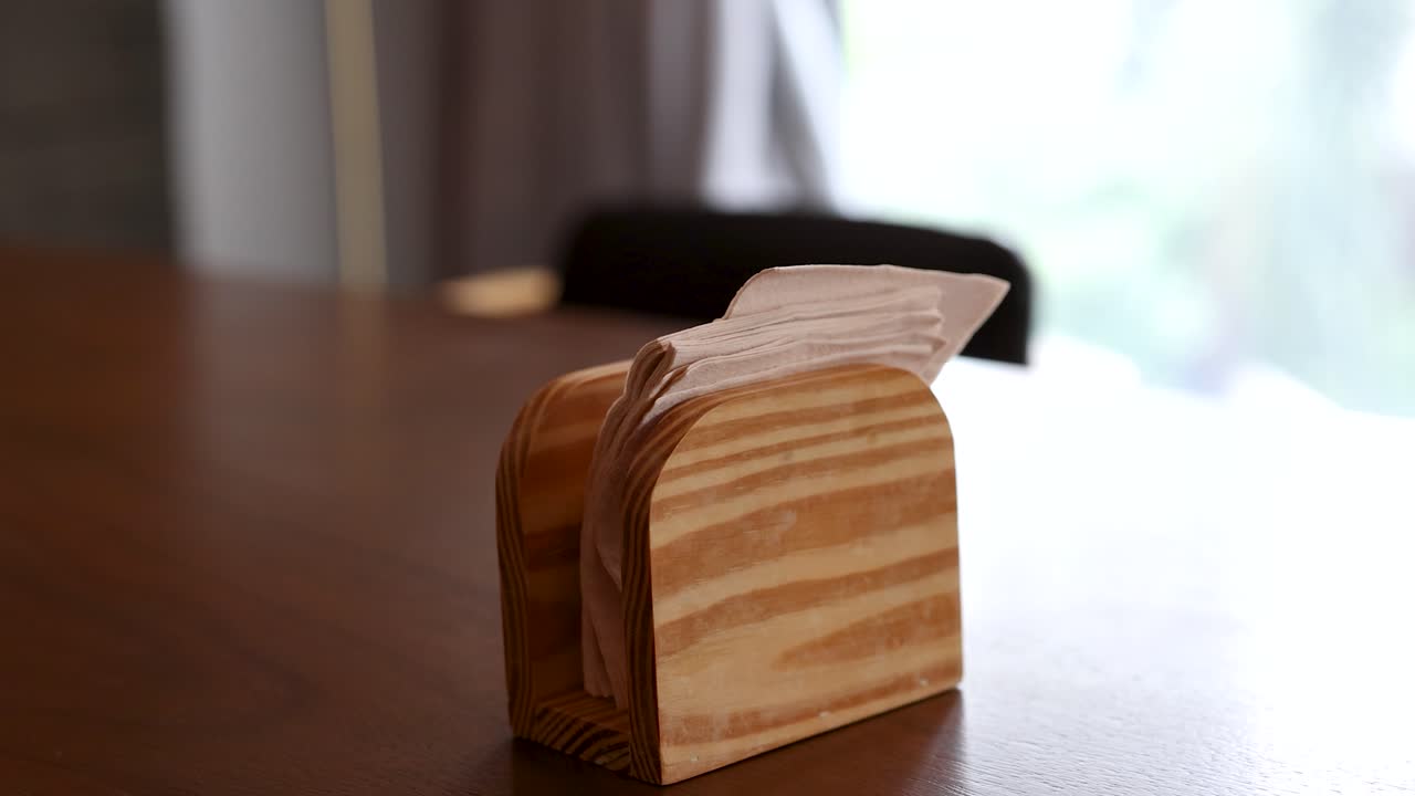 Hand reaches for napkin in wooden holder on table, natural light, casual dining setting