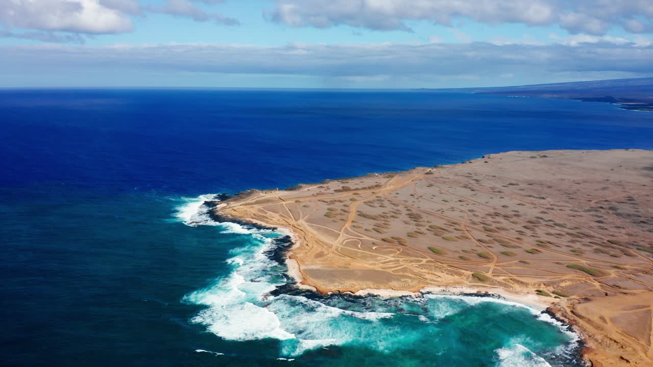 A rugged section of Hawaii’s southern coastline where turquoise waves strike volcanic rock and barren land crisscrossed by dusty trails, revealing the stark beauty of isolated tropical shores