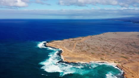 a rugged section of hawaii’s southern coastline where turquoise waves strike volcanic rock and barren land crisscrossed by dusty trails, revealing the stark beauty of isolated tropical shores