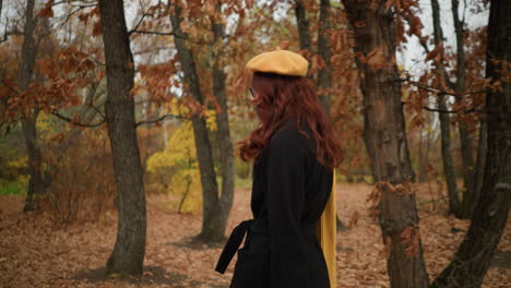 side view of lady strolling through forest, wearing yellow muffler, sunglasses, beret, and black coat, surrounded by autumn foliage, enjoying nature with a calm and relaxed expression