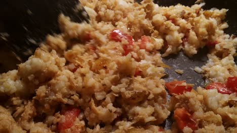 close up of stirring fried rice with tuna, tomatoes and cheese in a black plan with a wooden spatula