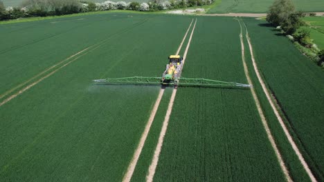 a drone shot following and descending towards a yellow and green tractor, spraying wheat fields with his machinery behind