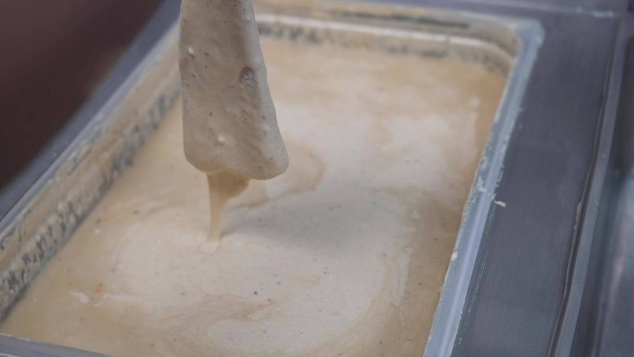 Gloved worker dips a food item into a deep tray of thick seasoned batter in a takeaway store kitchen.
