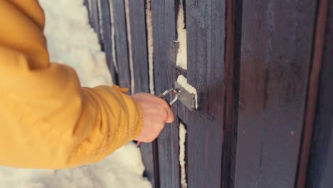 man unlocking frozen padlock of wooden gate in winter