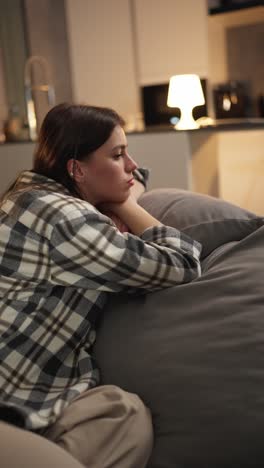 Vertical-video-of-a-tired-and-bored-brunette-girl-in-a-checkered-shirt-lies-on-the-sofa-leaning-on-the-back-and-on-the-pillow-in-a-modern-apartment-at-home-in-the-evening