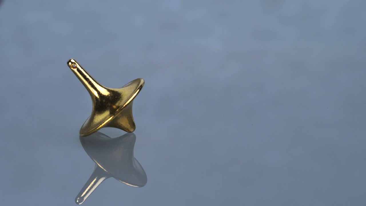 A golden, polished, spinning top wobbles on a reflective surface before stopping