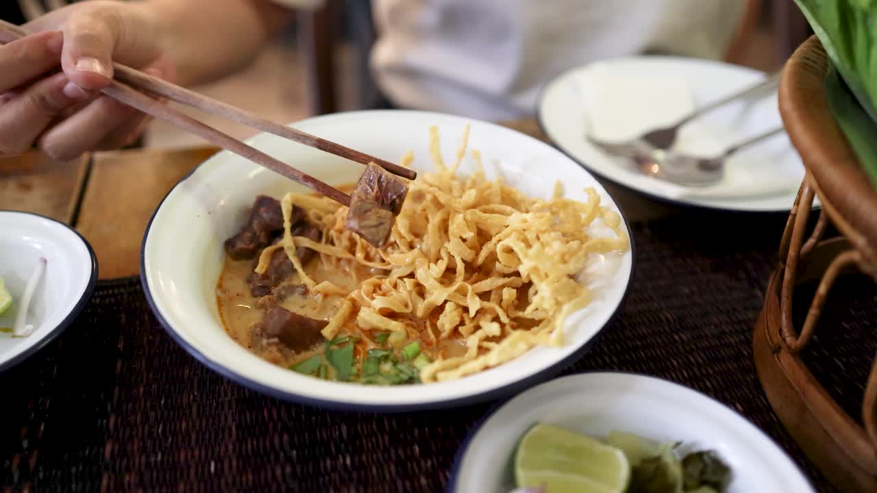 A person uses chopsticks to pick up a piece of beef from a bowl of Northern Thai curry noodles in a well-lit Bangkok restaurant, with close-up camera movement