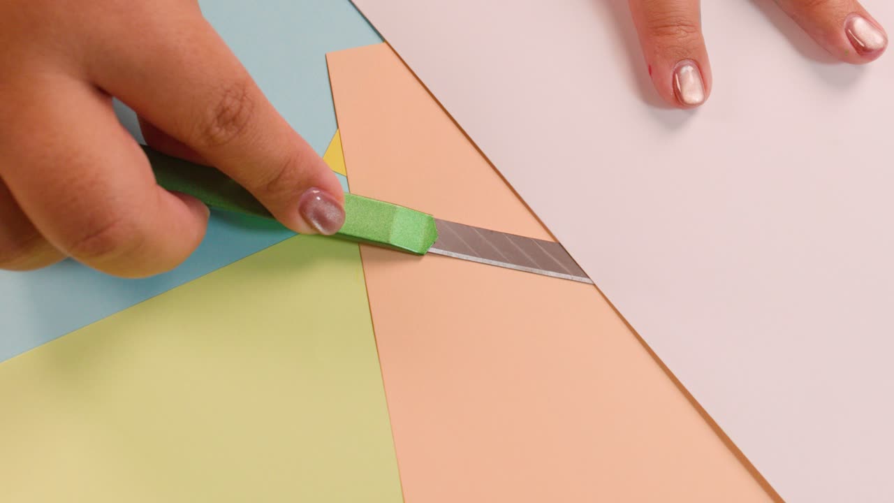 Hands use utility knife to cut colored paper on desk, bright lighting, overhead camera view