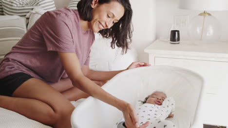 madre milenaria sentada cuidando a su bebé recién nacido durmiendo en una cuna junto a su cama