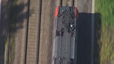 A-bird's-eye-drone-shot-captures-a-red-cabbed-diesel-locomotive,-with-its-gray-body-gleaming,-making-its-way-on-a-track-bedded-in-gravel,-flanked-by-a-grassy-embankment-and-a-sturdy-concrete-wall