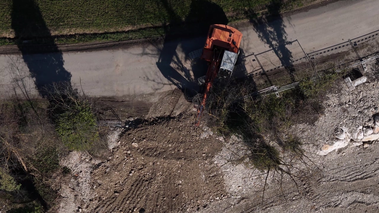 Aerial view of an orange excavator working on a demolition site, clearing rubble and debris near a road and grassy area