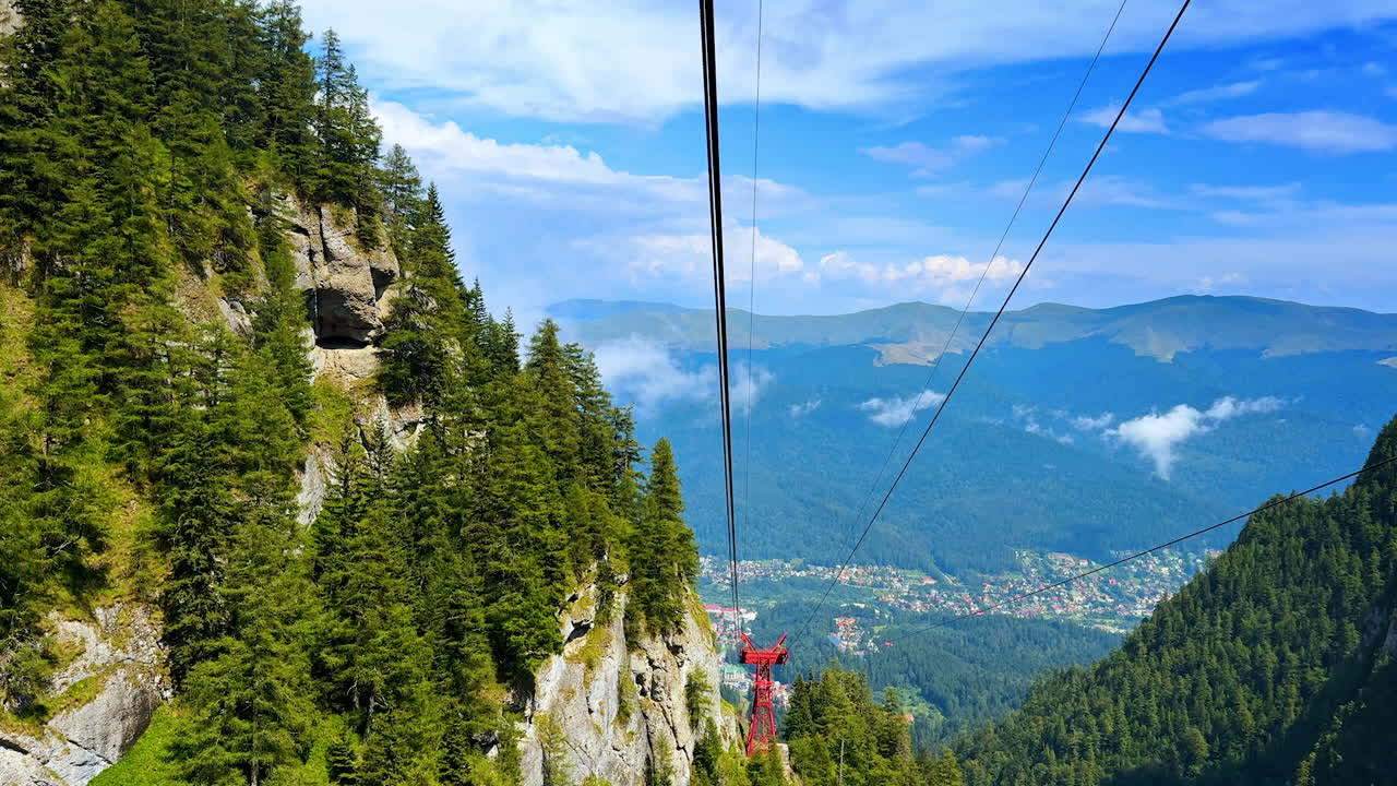 Una funivia viaggia sopra alberi alti e ripide scogliere. La vista mostra montagne distanti e una piccola città sottostante. Il cielo ha nuvole e tonalità blu, suggerendo una giornata limpida.