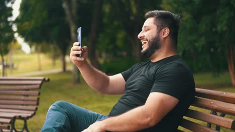joven con camisa negra sentado en un banco en el parque se comunica a través de un enlace de video en un turista en línea de teléfono móvil en una videollamada