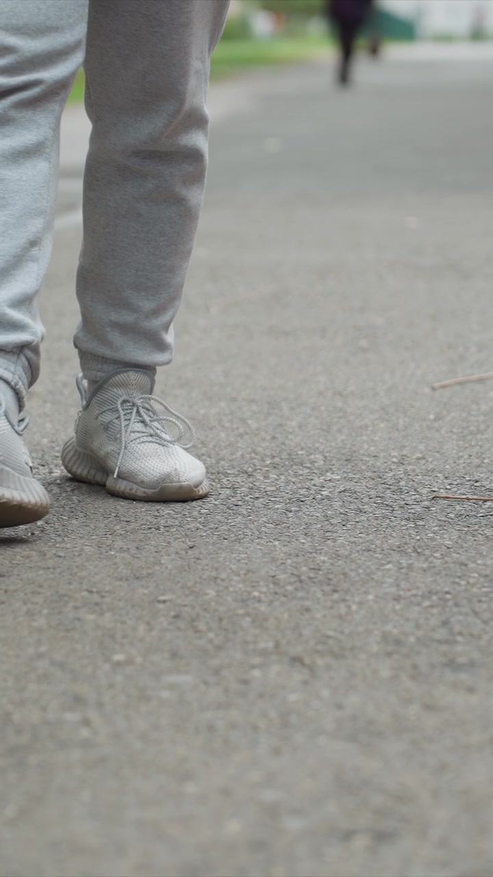 Man in light gray pants moving forward with steady steps. Gray pants and white shoes are prominent focus of camera. Outdoor setting with greenery and blurred distance background