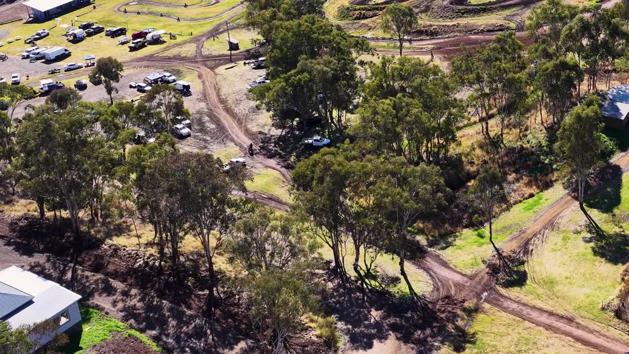 Drone captures dirt bike rider on winding forest trail in daylight, parked vehicles visible nearby