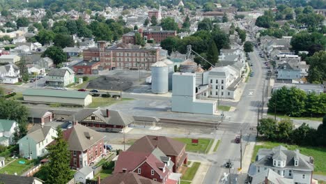 Rising-pullback-aerial-reveals-small-town-community-in-Midwest-USA,-rural-America,-houses-and-homes-line-street,-feed-mill,-businesses-and-traffic-on-street-below,-summer-daytime-establishing-shot