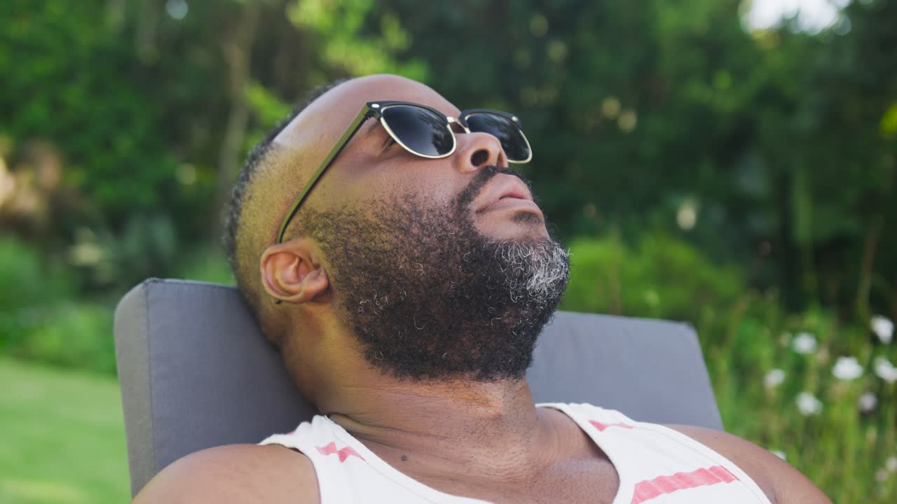 African American mid-adult man nodding reclining on chair in garden in shades striped tank settling