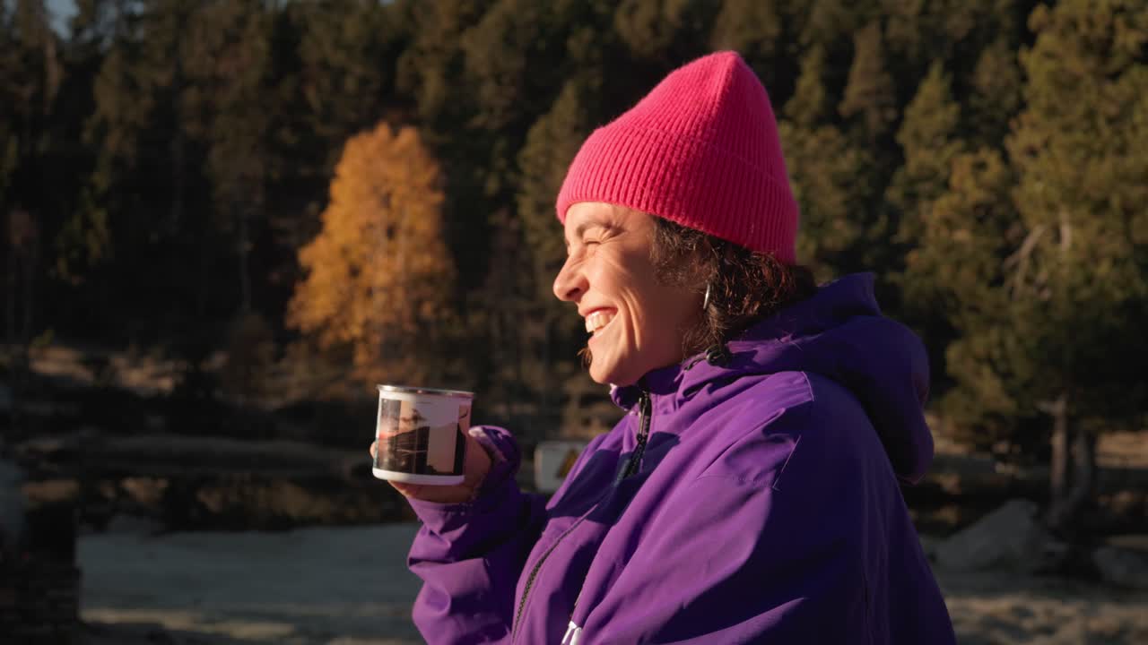 Slow-motion side view of a joyful woman wearing a winter cap, laughing and sipping coffee outdoors in the mountains during winter morning.