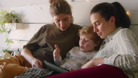 kid using tablet and chatting with lesbian mothers in bedroom