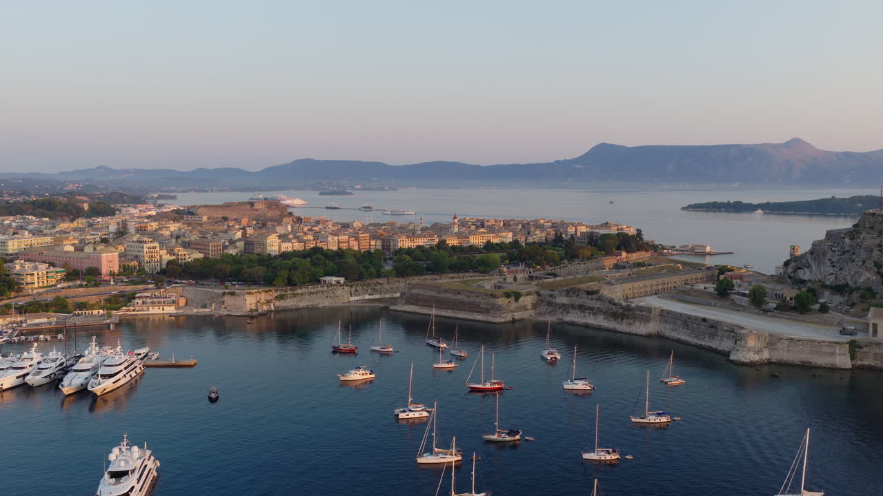 Old town of Corfu at sunrise, rooftops and harbor visible under golden morning light, slow approach over sailboats at dawn