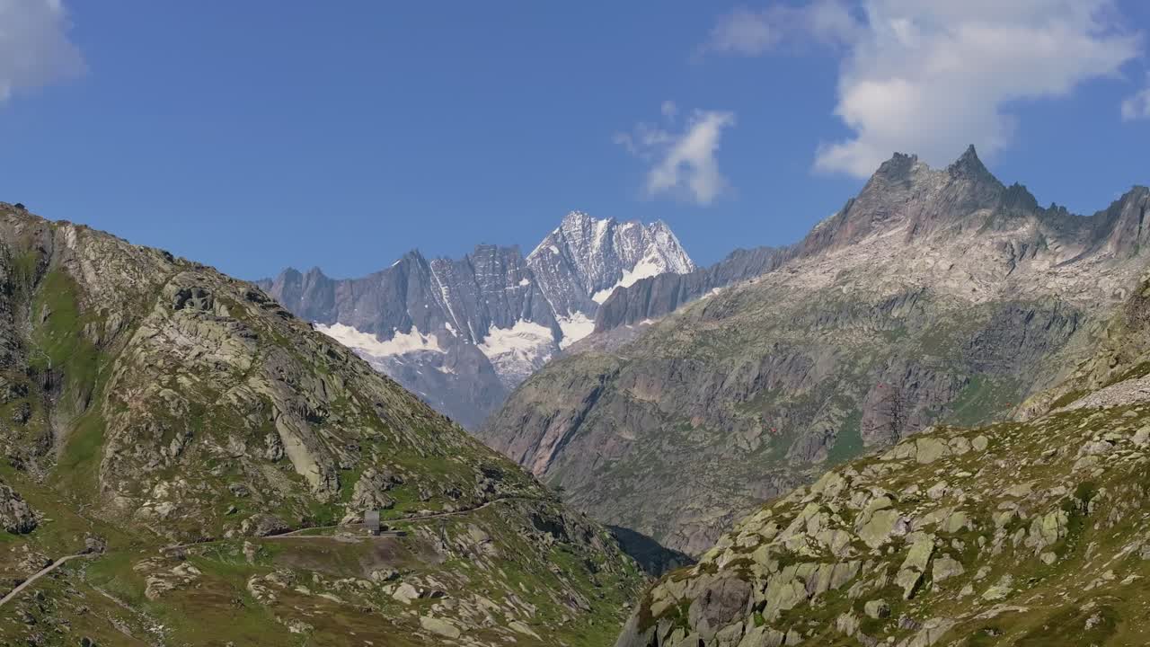 Revealing drone shot showing a breathtaking mountain landscape with rugged rocky slopes, alpine meadows, and snow-capped peaks under a clear blue sky