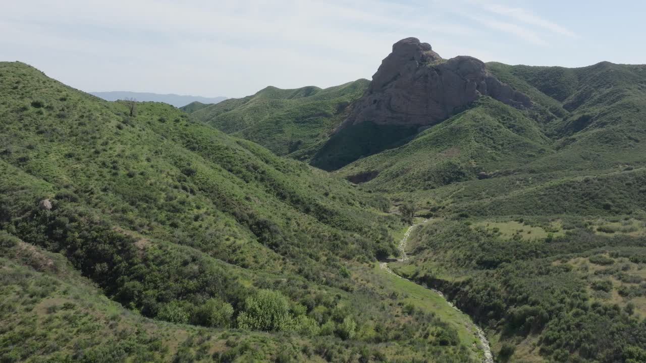 Aerial push-in shot moving toward a large rock monument among green hills covered in vegetation with a blue partly cloudy sky.