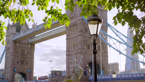 Tower-Bridge-from-the-river-Thames-side-in-London,-beautiful-trees-and-lamppost-with-red-double-decker-bus-passing-by,-on-a-sunny-summer-day-with-flares