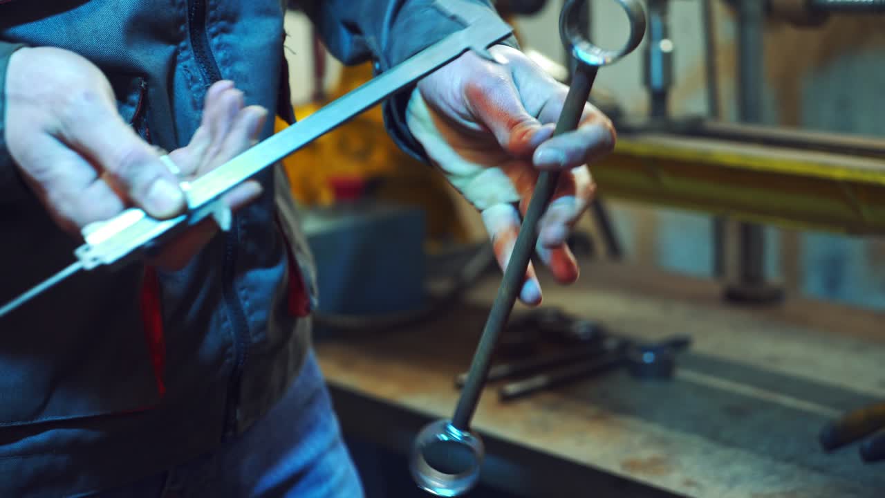 Mechanic is working with metal in a workshop, making measurements of metal parts. Worker holds a caliper in his hand, in a special suit on factory background.