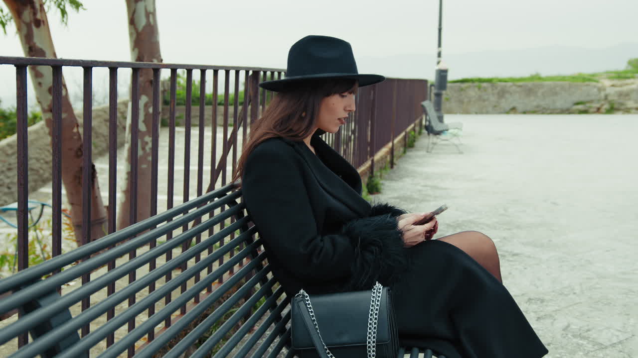 Elegant Woman With A Black Hat Using The Smartphone While Sitting On The Bench