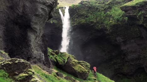 Tomas-En-Cámara-Lenta-De-Una-Remota-Cascada-Islandesa-Con-Un-Hombre-Caminando-Hacia-Ella