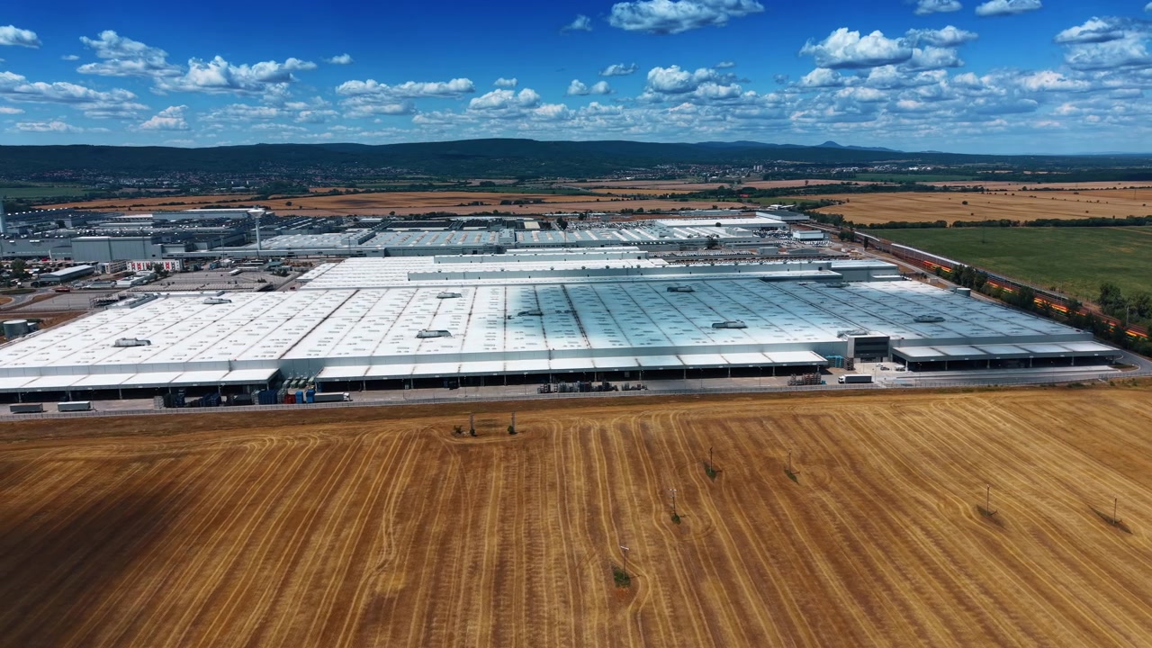 Grande complesso industriale accanto a campi mietuti. Tetti bianchi di fabbriche si estendono su terreni agricoli con binari ferroviari, colline e nuvole sparse all'orizzonte