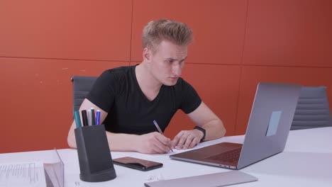 un joven europeo trabaja usando una computadora portátil, firma documentos con un bolígrafo, trabaja en línea en una oficina roja brillante