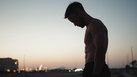 Silhouette-Eines-Muskulösen-Bodybuilders-Mit-Perfektem-Körper,-Der-Bei-Sonnenaufgang-Oder-Sonnenuntergang-In-Einem-Fitnessstudio-Am-Strand-Posiert