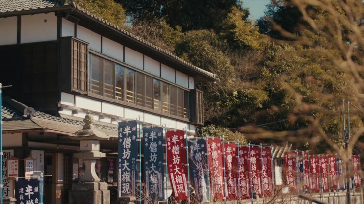 L'edificio tradizionale del santuario in legno di Hansobo, situato in cima al complesso del tempio Kencho-ji a Kamakura, incorniciato da file di stendardi cerimoniali e da una collina boscosa, con una calda luce autunnale sugli alberi