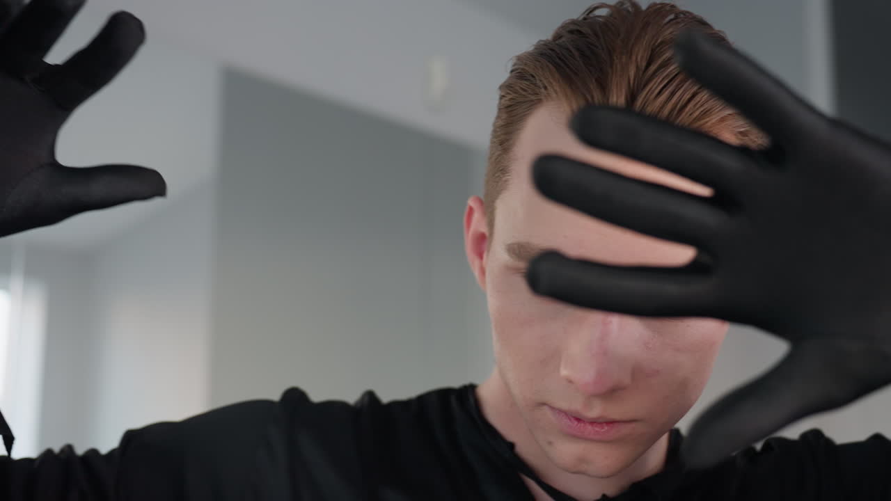 close up of young man in black outfit playfully interacting with camera, moving gloved hand across lens, revealing focused gaze and subtle smile in bright modern mirror studio setting