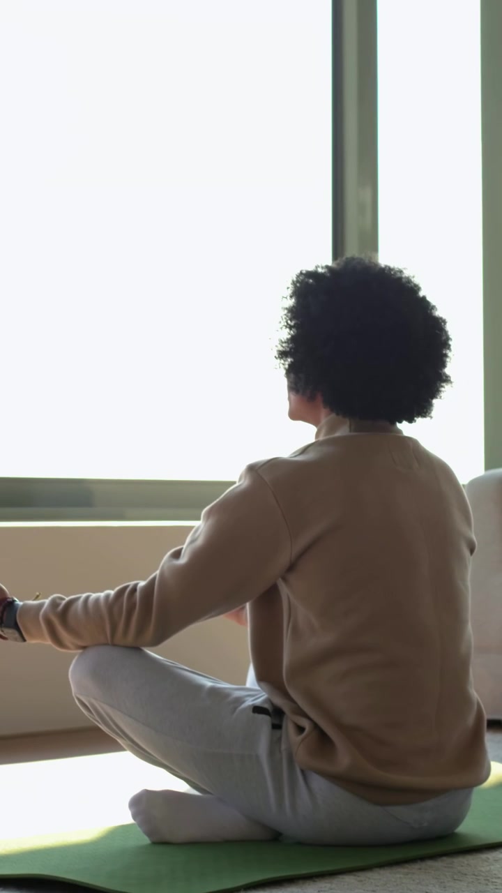 Video verticale: Un uomo adulto che indossa un maglione color cammello entra e srotola un tappetino verde a casa per meditare.