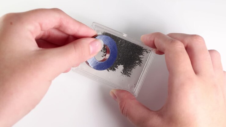 Hands manipulate a ring magnet over iron filings, demonstrating magnetic attraction and repulsion on a white background