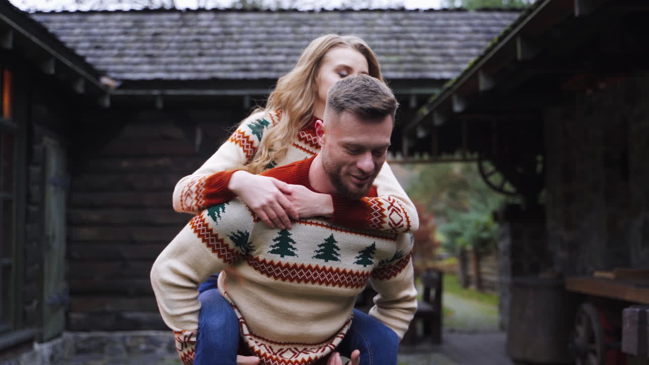 Happy young lovely couple having fun near wooden house in the park. Man piggybacking girl, running happily. Guy whirling woman around. Love story video.