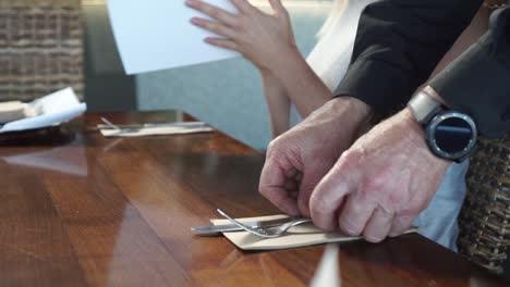 Slowmo---Männlicher-Kellner-Legt-Besteck-Auf-Holztisch,-Während-Junge-Frau-Mit-Blonden-Haaren-Die-Speisekarte-In-Einem-Luxusrestaurant-Betrachtet---Niedrige-Nahaufnahme