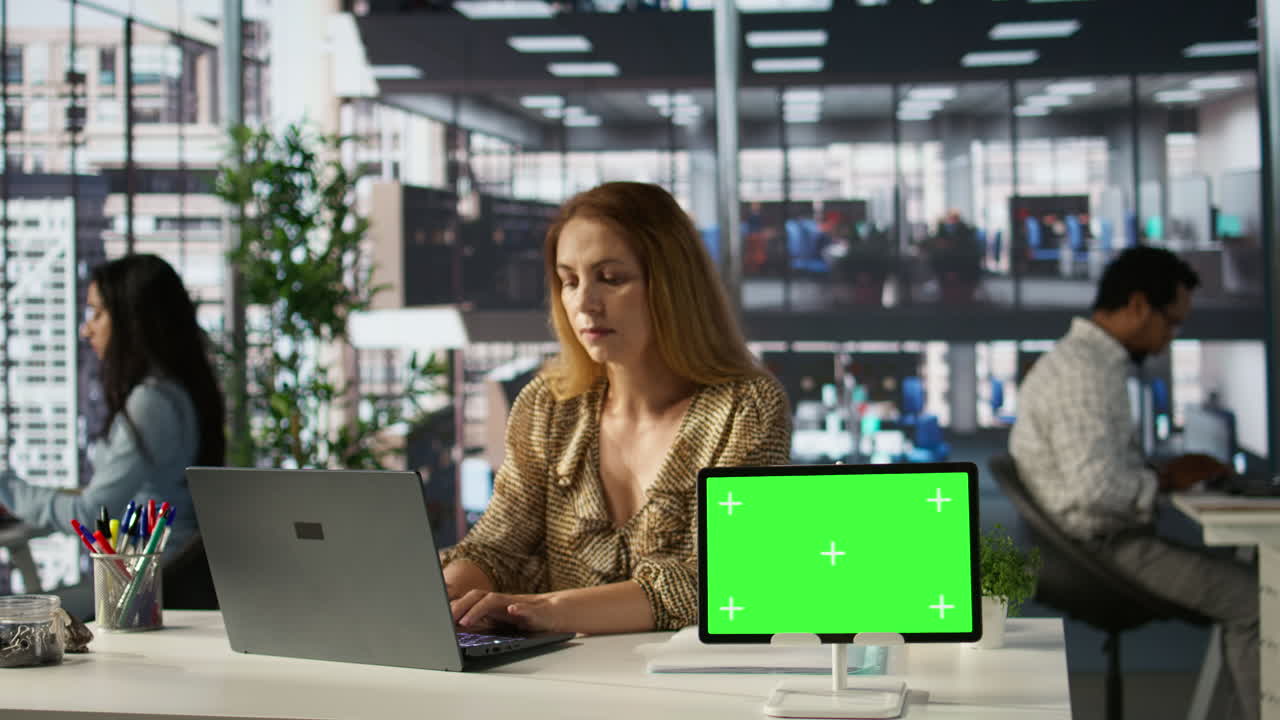 Woman working on laptop with green screen tablet in office