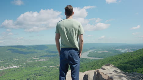 man standing on top of mountain looking at amazing epic view of green hills and river