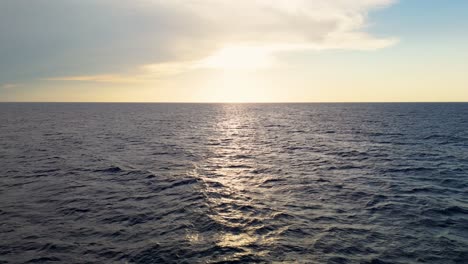 peaceful aerial dolly out to setting sun with cascading ray of golden light over dark blue waters below cloudy sky