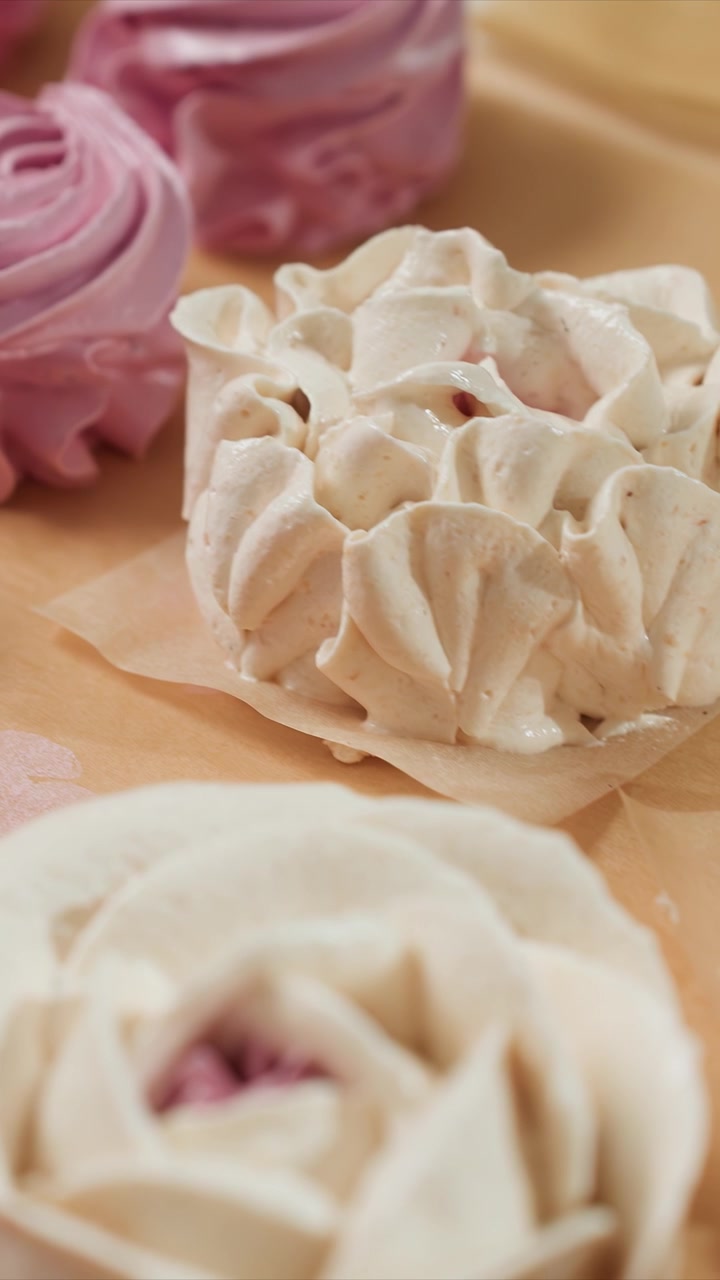 Close-up meringue berbentuk mawar merah muda dan putih yang tersusun rapi. Kamera bergerak mulus untuk memperlihatkan detail seperti tepi bergelombang. Latar belakang menampilkan pola bunga samar yang menonjolkan