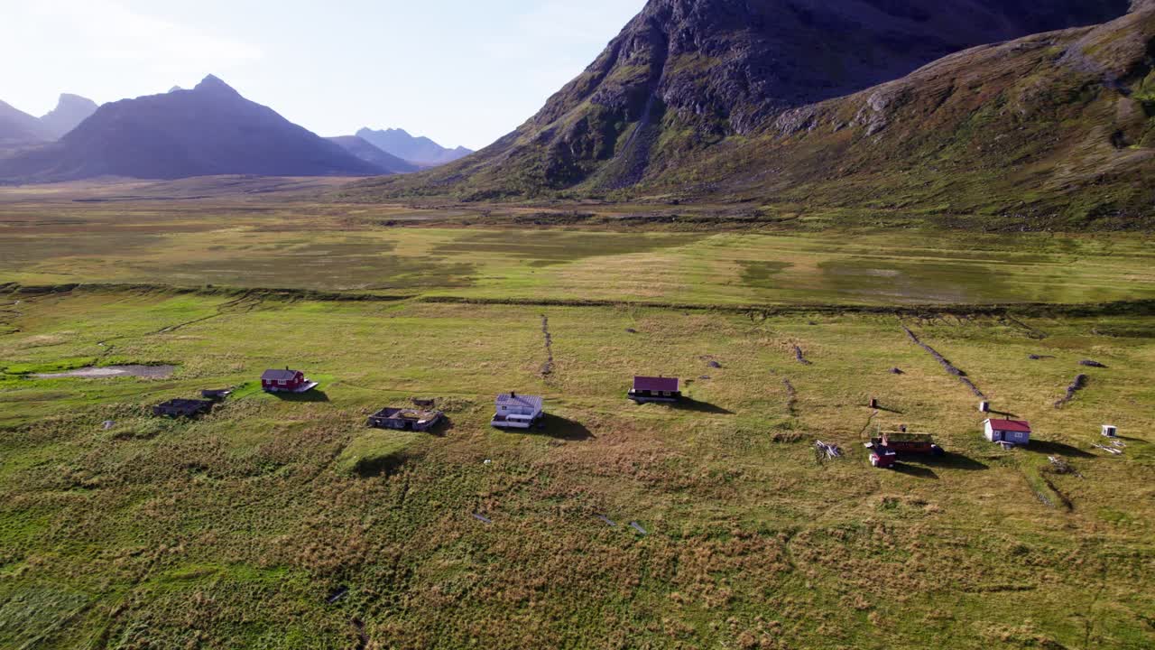 Flygfilmsklipp med drönare som flyger från höger till vänster över utspridda hus på gröna fält mellan berg, med respektingivande berg i bakgrunden, ett cinematiskt nordiskt landskap.