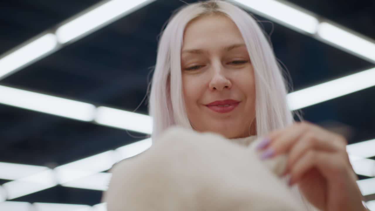 Portrait of white long haired woman smiling while admiring toddler hoodie in retail store holding plush fabric with manicured nails exploring texture and hood detail against blurred racks