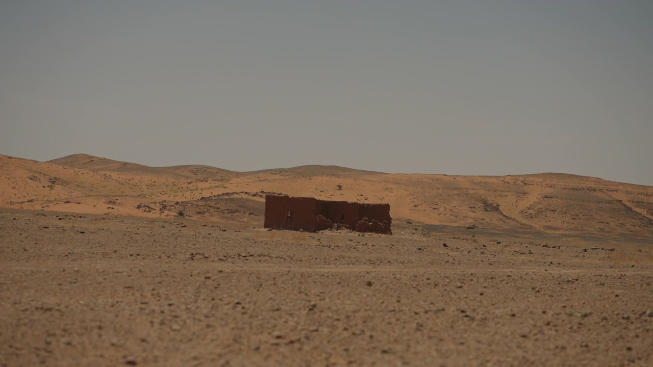 Capanna solitaria in mattoni di fango che si erge nella vasta pianura desertica del Sahara sotto un cielo velato nel Marocco meridionale.