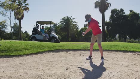 Two-caucasian-women-playing-golf-wearing-face-masks-one-taking-shot-from-bunker